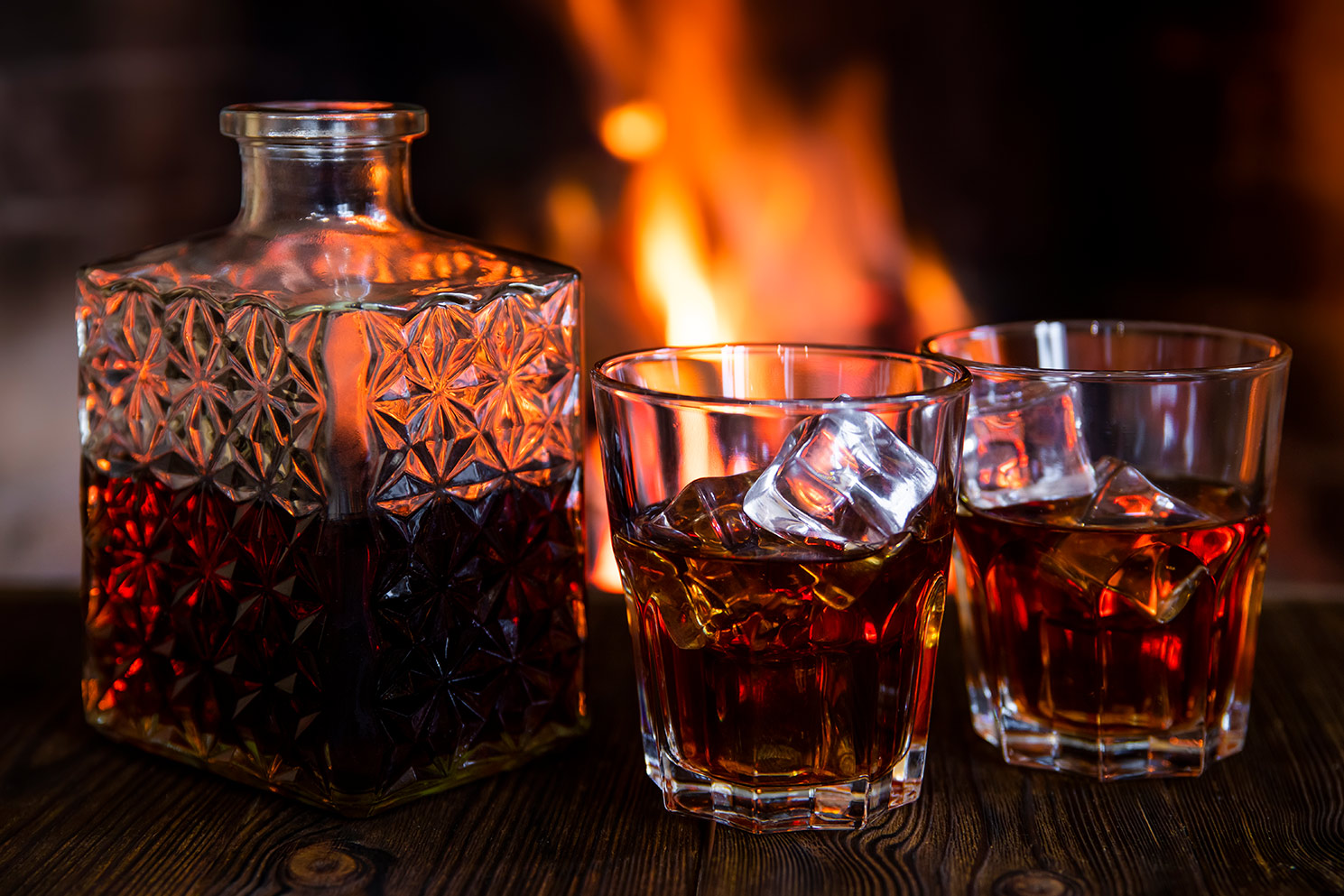 A decanter of whiskey and two glasses with ice on wood table in front of a bonfire or fireplace