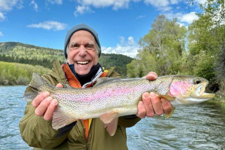 Famous actor, Henry Winkler, stands holding a large fish in a hat and many jackets on the water