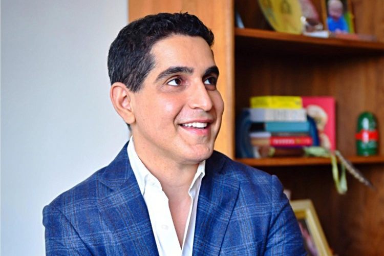 Man smiles in suit with bookshelf behind